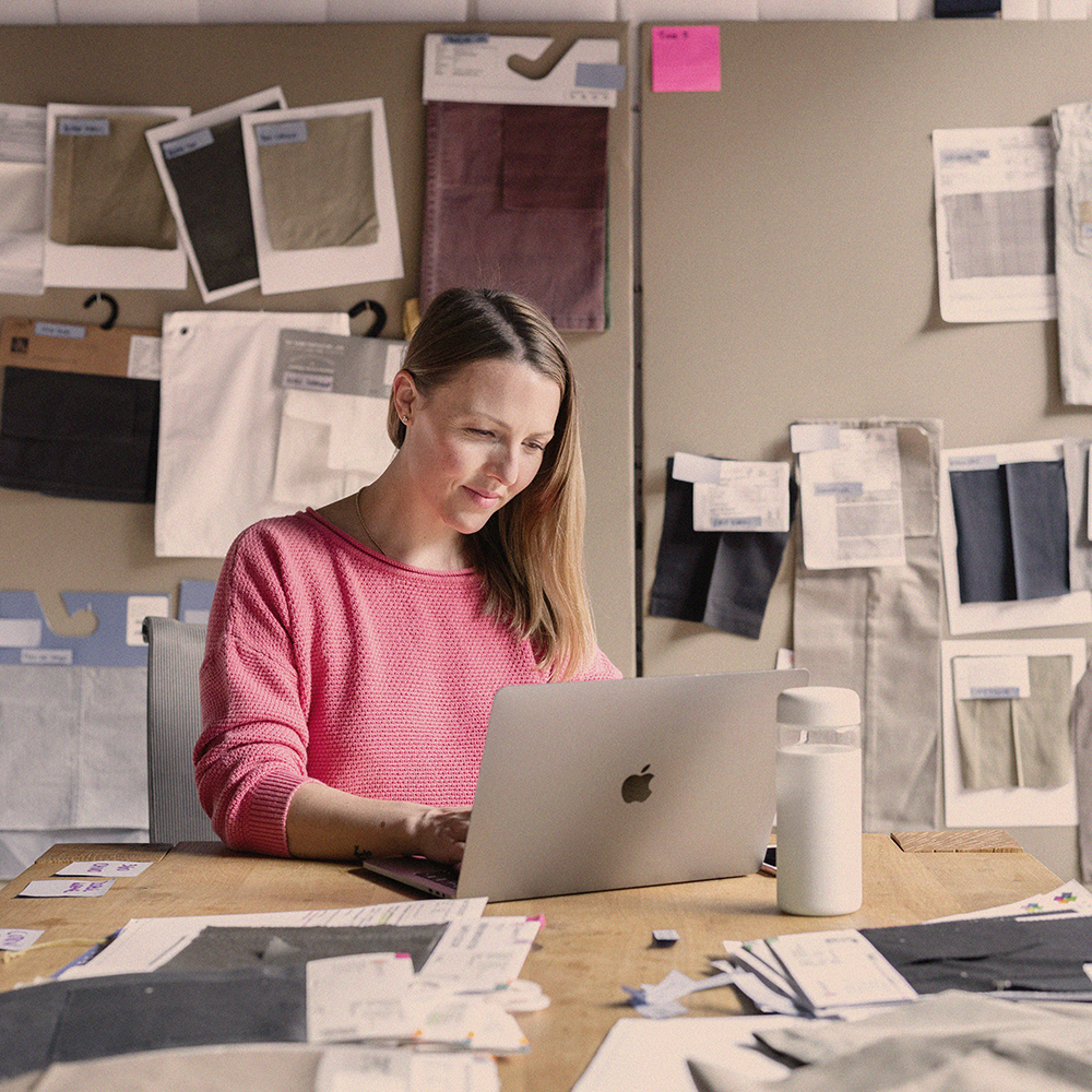 A business woman sits at her laptop in her work room, surrounded by papers and samples.